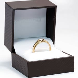 Pode incluir: Um anel de ouro com uma configura&ccedil;&atilde;o de diamante em uma caixa de anel marrom.