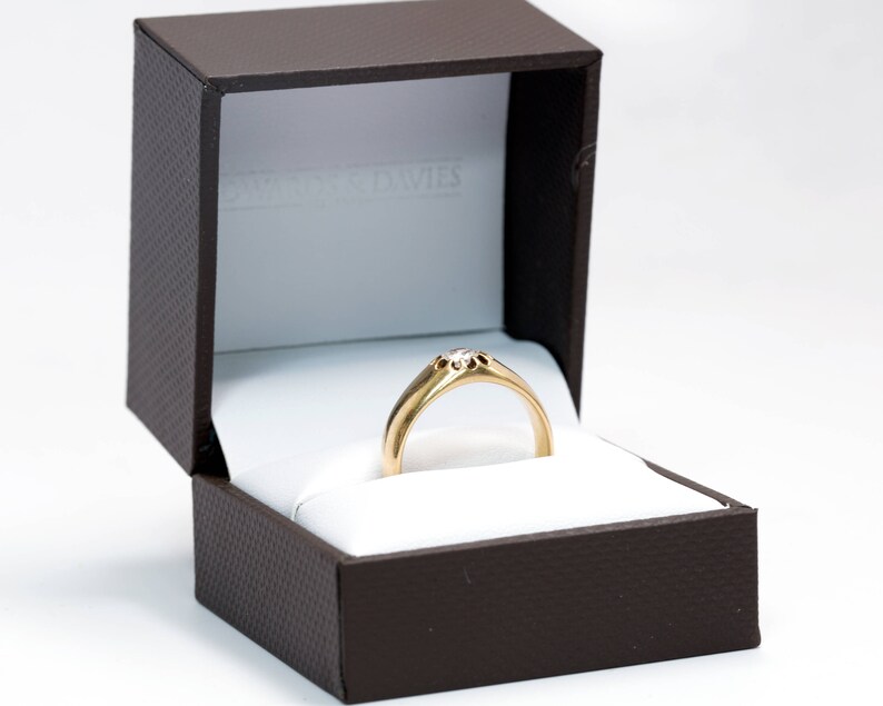 Pode incluir: Um anel de ouro com uma configura&ccedil;&atilde;o de diamante em uma caixa de anel marrom.