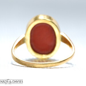 May include: A gold ring with a red oval-shaped gemstone set in a bezel setting. The ring is shown from the back, revealing the smooth gold band.