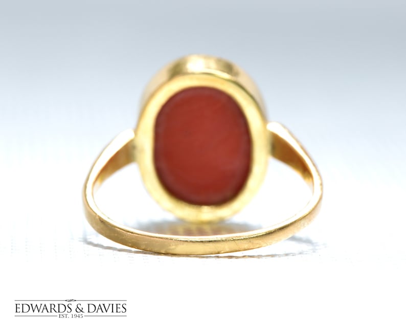 May include: A gold ring with a red oval-shaped gemstone set in a bezel setting. The ring is shown from the back, revealing the smooth gold band.