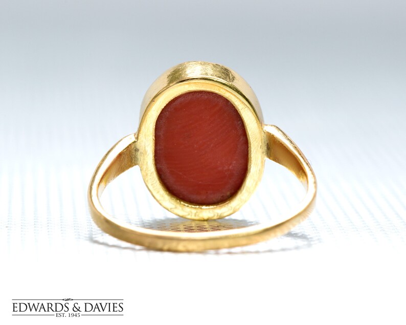 May include: A gold ring with a red oval-shaped gemstone. The ring is viewed from the back, showing the setting of the stone.