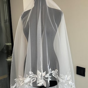 May include: A white bridal veil with a delicate floral pattern around the edge. The veil is made of soft tulle and is perfect for a wedding.