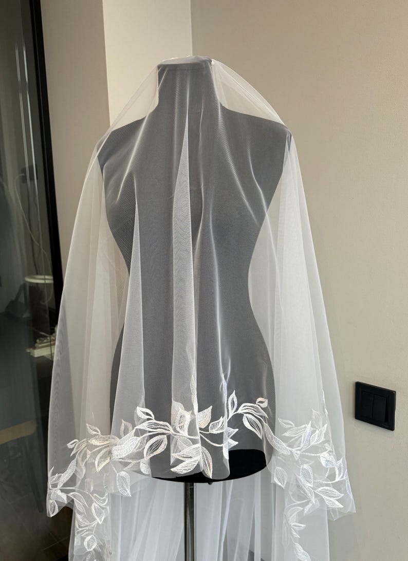 May include: A white bridal veil with a delicate floral pattern around the edge. The veil is made of soft tulle and is perfect for a wedding.