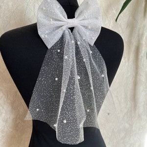 May include: A white tulle bow with scattered faux pearls, displayed on a black mannequin. The bow is large and airy, with long, flowing tails. The tulle has a delicate, dotted pattern, creating a soft, elegant look. This accessory is suitable for weddings or special occasions.