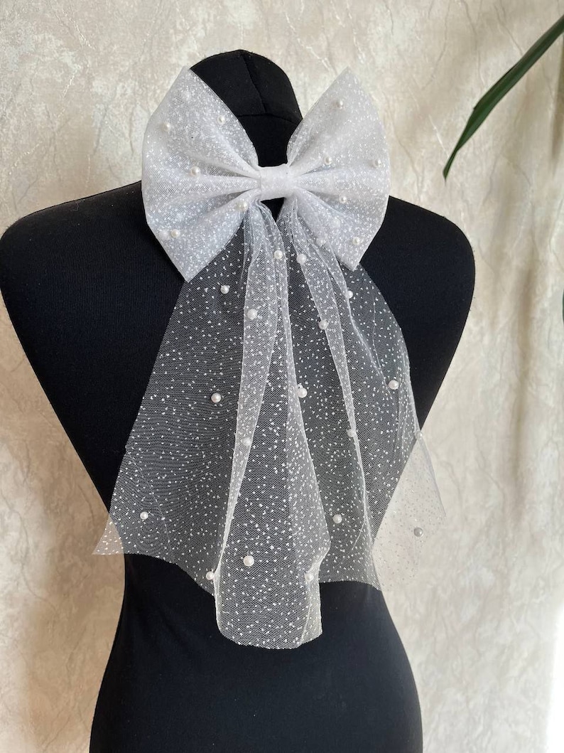 May include: A white tulle bow with scattered faux pearls, displayed on a black mannequin. The bow is large and airy, with long, flowing tails. The tulle has a delicate, dotted pattern, creating a soft, elegant look. This accessory is suitable for weddings or special occasions.