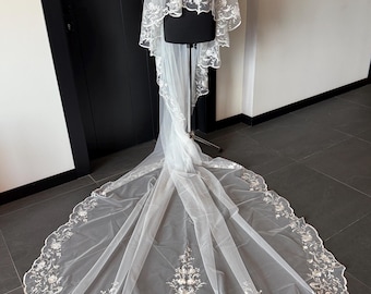 Puede incluir: Un velo de novia blanco con intrincados bordados florales. El velo se exhibe sobre un maniquí, mostrando su delicado diseño y larga cola. El bordado se concentra a lo largo de los bordes y la cola.