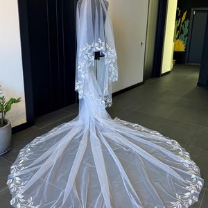 May include: A white bridal veil with a delicate floral pattern around the edges. The veil is made of soft tulle and is long enough to flow behind the bride as she walks down the aisle.