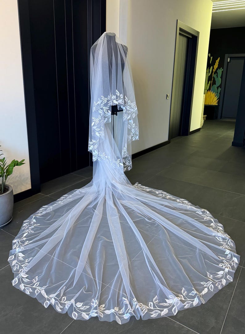 May include: A white bridal veil with a delicate floral pattern around the edges. The veil is made of soft tulle and is long enough to flow behind the bride as she walks down the aisle.