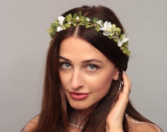 May include: A floral crown featuring white flowers and green leaves. The crown is worn on the head and is made of artificial flowers. The woman in the image is wearing a white lace top.