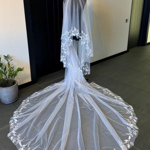 May include: A white bridal veil with a delicate floral design. The veil is made of soft tulle and features a scalloped edge. The floral design is embroidered in a subtle white thread.