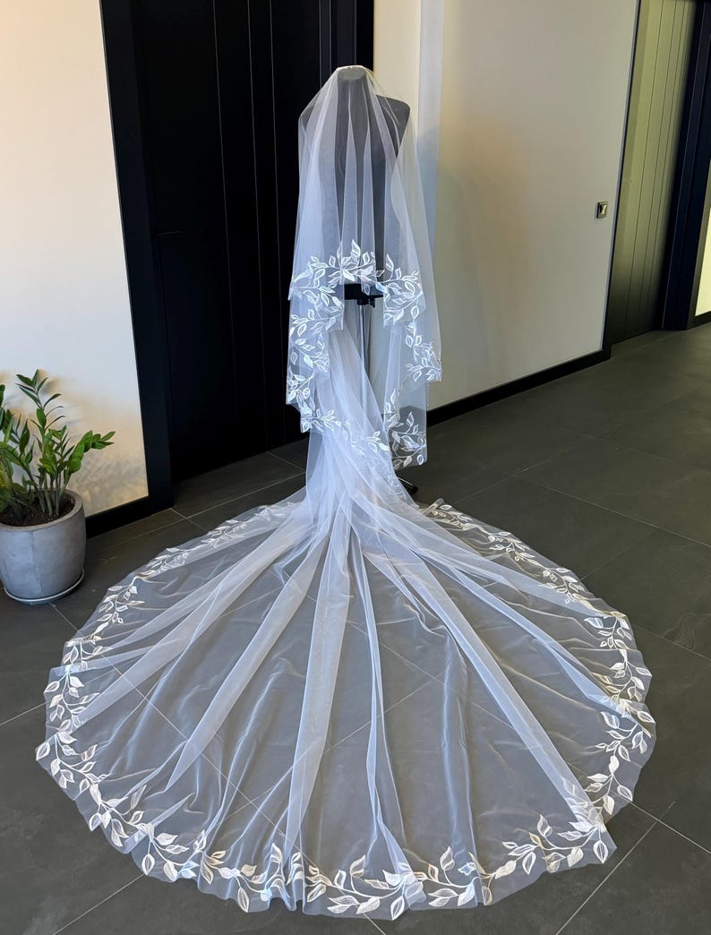 May include: A white bridal veil with a delicate floral design. The veil is made of soft tulle and features a scalloped edge. The floral design is embroidered in a subtle white thread.