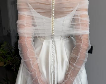 May include: A white off-the-shoulder wedding dress with a tulle overlay and long sleeves. The dress has a fitted bodice and a flowing skirt. The tulle overlay is gathered at the shoulders and the sleeves are gathered at the wrists. The dress is fastened with pearl buttons down the back.