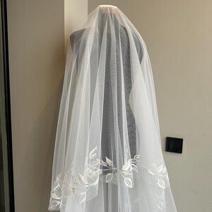 May include: A white bridal veil with a delicate floral pattern embroidered around the edge. The veil is made of soft tulle and is designed to be worn over the head.