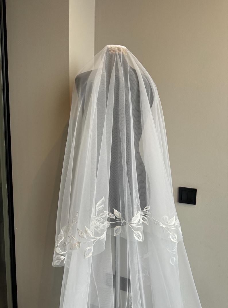May include: A white bridal veil with a delicate floral pattern embroidered around the edge. The veil is made of soft tulle and is designed to be worn over the head.