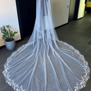 May include: A white tulle wedding veil with a delicate floral embroidery design around the edge.