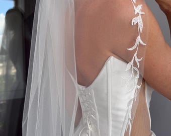 May include: A white wedding dress with a corset bodice and a flowing skirt. A sheer veil with embroidered floral detailing drapes over the shoulder. The dress features a lace-up back and a classic, elegant design.
