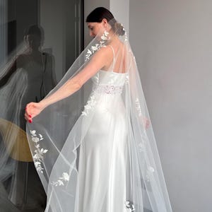 Pode incluir: Um vestido de noiva branco com um véu longo e esvoaçante. O véu é adornado com delicados bordados florais nas bordas. O vestido apresenta um design simples e elegante com alças finas e um detalhe de renda na cintura.