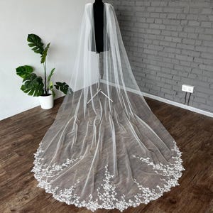 May include: A long, white bridal veil with a delicate floral lace trim. The veil is displayed on a black mannequin, showcasing its flowing design. The lace trim features intricate floral patterns.