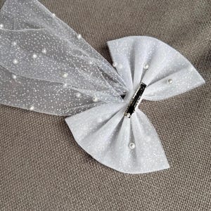 May include: A white glitter bow with a sheer, dotted veil. The bow is adorned with small, faux pearls and a silver clip. This hair accessory is suitable for special occasions.