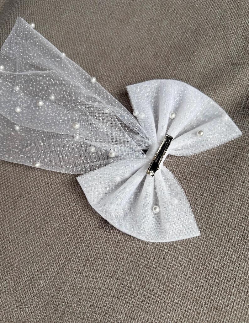 May include: A white glitter bow with a sheer, dotted veil. The bow is adorned with small, faux pearls and a silver clip. This hair accessory is suitable for special occasions.