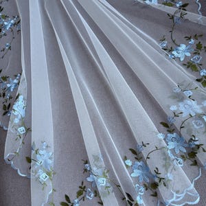 May include: A white, sheer veil with blue floral embroidery along the scalloped edge. The embroidery features light blue flowers and green leaves. The veil is made of a lightweight, flowing fabric, suitable for a wedding or special occasion.