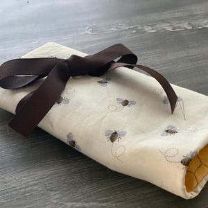 May include: A white fabric pouch with a brown ribbon tied around it. The pouch is decorated with a pattern of small bees and has a yellow honeycomb lining.