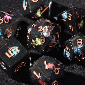 Peut inclure: Ensemble de sept dés polyédriques noirs avec des chiffres cuivrés et des motifs de papillons. Les dés sont destinés aux jeux de rôle et comprennent un d4, d6, d8, d10, d12, d20 et un dé de pourcentage.