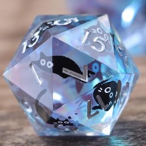 Puede incluir: Un dado D20 azul claro multifacético con ilustraciones de gatos negros y numeración blanca. El dado tiene una calidad brillante e iridiscente, con inclusiones visibles. Los números están en una fuente serif.