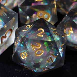 Puede incluir: Juego de dados transparentes e iridiscentes con números dorados. Los dados son de múltiples caras, incluido un dado de veinte caras. Los dados tienen una apariencia brillante, similar a una galaxia, con colores azul, verde y morado.