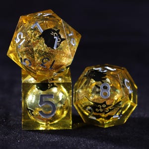 May include: Three yellow resin dice with silver numbering and black cat illustrations. The dice are a cube, a 20-sided die, and a 12-sided die. The dice have gold glitter suspended within the resin.