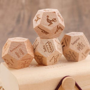 Può includere: Quattro dadi dodecaedrici in legno con immagini e parole incise come "Romantico", "Subs" e "Sorpresa". I dadi sono impilati su una scatola di legno, con la data "10.12.2024" visibile.
