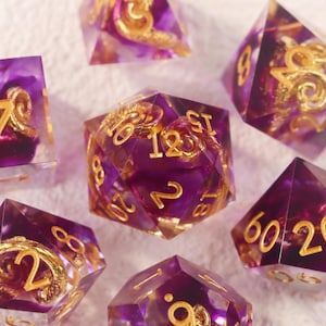 May include: Close-up of a set of polyhedral dice. The dice are a translucent purple with gold-colored numbers and decorative swirls inside. The dice are designed for tabletop role-playing games.