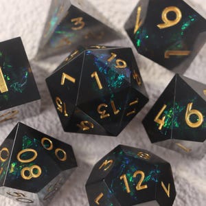 Peut inclure: Un ensemble de dés polyédriques noirs avec des inclusions irisées vertes et bleues. Les chiffres dorés ressortent sur les différentes formes, dont un D20. L'ensemble est présenté sur une surface claire.