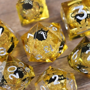 Peut inclure: Un ensemble de dés dorés remplis de paillettes, avec des chiffres blancs et des silhouettes de chats noirs. Les dés sont de formes variées, dont un dé à vingt faces, posés sur une surface en bois.