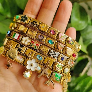 Puede incluir: Pulseras de dijes doradas con varios dijes decorativos, incluyendo corazones, flores y formas geométricas. Algunos dijes tienen piedras preciosas de colores. Las pulseras se sostienen en una mano.