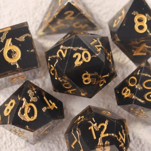 Op de afbeelding: Een verzameling zwarte polyhedrale dobbelstenen met gouden cijfers en details. De dobbelstenen hebben verschillende geometrische vormen, perfect voor rollenspellen en bordspellen.
