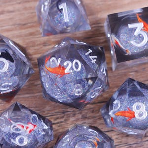 Puede incluir: Primer plano de varios dados transparentes de múltiples caras con números blancos y detalles en naranja. Los dados están llenos de purpurina azul. Los dados están colocados sobre una superficie de madera marrón claro.