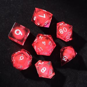 Pode incluir: Conjunto de sete dados poliédricos vermelhos translúcidos com numeração prateada. Os dados estão dispostos sobre uma superfície preta. O conjunto inclui um dado de quatro lados, seis lados, oito lados, dez lados, doze lados, vinte lados e um dado percentual.