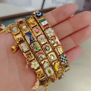 Può includere: Una collezione di bracciali a maglie modulari color oro con vari ciondoli in smalto colorato, tra cui un occhio greco, una bandiera messicana, fiori, una banana, un motivo a scacchi e la parola Love, con ciondoli pendenti a forma di farfalla.