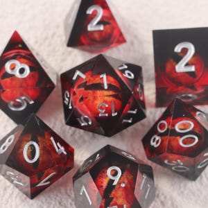 May include: A set of polyhedral dice with a fiery eye design. The dice are black with red and orange accents, and silver numbering. The set includes various shapes, such as d4, d6, d8, d10, d12, and d20, perfect for tabletop gaming.
