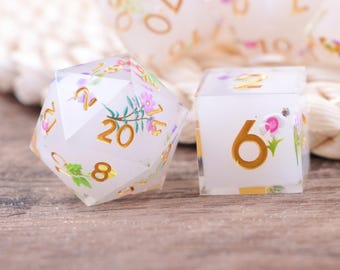 May include: A set of clear, floral-themed dice. The dice feature gold numbering and are adorned with colorful flowers and greenery. The image shows a twenty-sided die and a six-sided die, with other dice visible in the background.