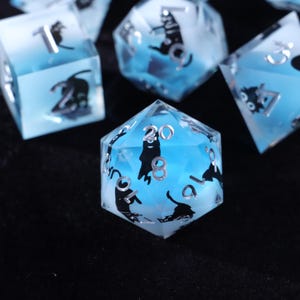 Puede incluir: Un conjunto de dados poliédricos con un diseño translúcido azul y blanco. Cada dado presenta números plateados y siluetas de gatos negros. Los dados están diseñados para juegos de mesa.