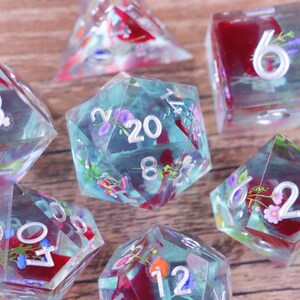 Peut inclure: Un ensemble de dés en résine transparente avec des inclusions florales. Les dés présentent diverses formes, dont un dé à vingt faces, et sont remplis de fleurs colorées et d'accents rouges. Les chiffres sont visibles sur chaque dé.