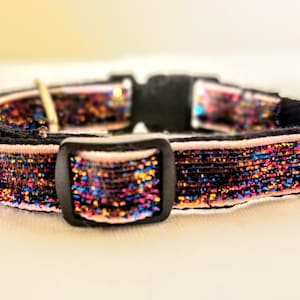 May include: A pink and black cat collar with a sparkly iridescent ribbon. The collar has a black plastic buckle.