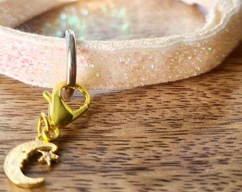 May include: A gold-colored charm with a crescent moon and star pendant hangs from a silver clasp. The charm is attached to a glittery, light pink bracelet. The bracelet is resting on a wooden surface.