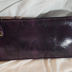 May include: A rectangular dark purple clutch bag with a glossy snakeskin-like texture. The bag features a gold-tone metal turn-lock closure on the left side, attached to a small tab. The surface has a reflective finish and a consistent scale pattern.