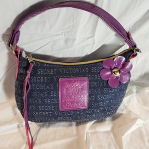 May include: A dark blue denim handbag featuring a repeating Victoria's Secret logo print. It has a purple handle, a purple flower accent with a gold center, and a central purple patch with the text With LOVE Victoria's Secret in pink.