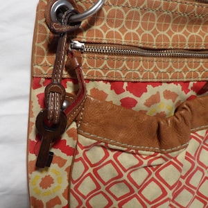 May include: A canvas crossbody bag featuring a geometric pattern in orange, red, and cream. The bag includes a brown leather pocket, a silver-toned metal zipper, and a brown leather key-shaped charm attached to a silver ring on the side strap.