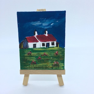 May include: A small painting on a wooden easel depicts a white cottage with a red roof against a blue sky. The cottage is surrounded by green grass and red flowers.