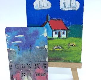 Könnte beinhalten: Zwei Gemälde auf Leinwand, eines zeigt ein Haus mit rotem Dach und blauem Himmel mit weißen Wolken, das andere zeigt eine Reihe von Häusern mit lila und blauem Himmel. Das Gemälde mit dem Haus steht auf einem Staffelei.